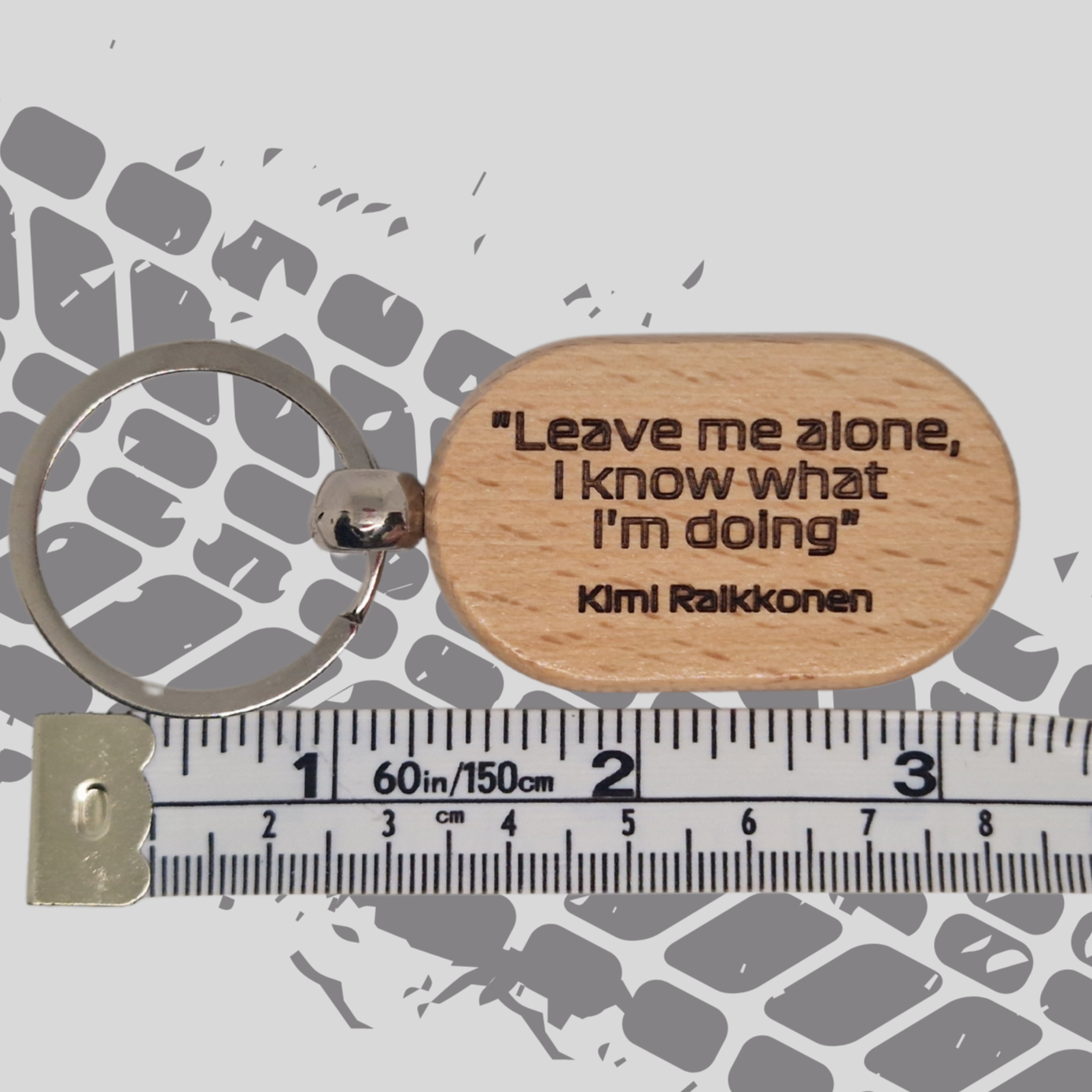 A wooden keychain engraved with the quote "Leave me alone, I know what I'm doing," attributed to Kimi Räikkönen, placed next to a tape measure for size comparison. The keychain’s natural wood grain is visible, with the bold engraving on the front. The tape measure highlights the keychain's size.