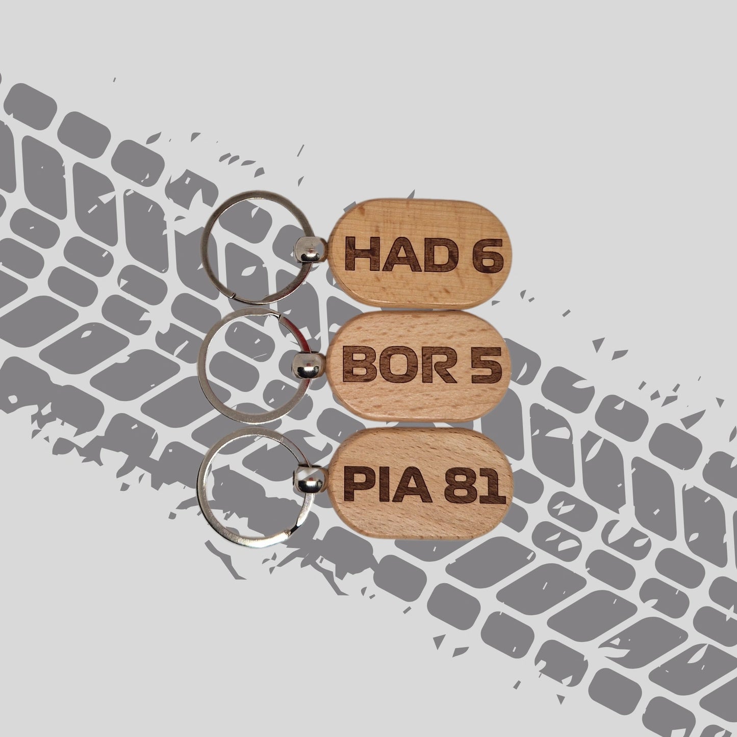 Three wooden keychains featuring F1 driver abbreviations and numbers for the 2025 season, displayed against a tyre tread background. Each keychain has a unique design with the driver's initials and number engraved on the wood