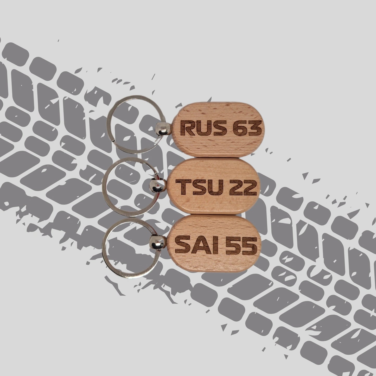 Three wooden keychains featuring F1 driver abbreviations and numbers for the 2025 season, displayed against a tyre tread background. Each keychain has a unique design with the driver's initials and number engraved on the wood