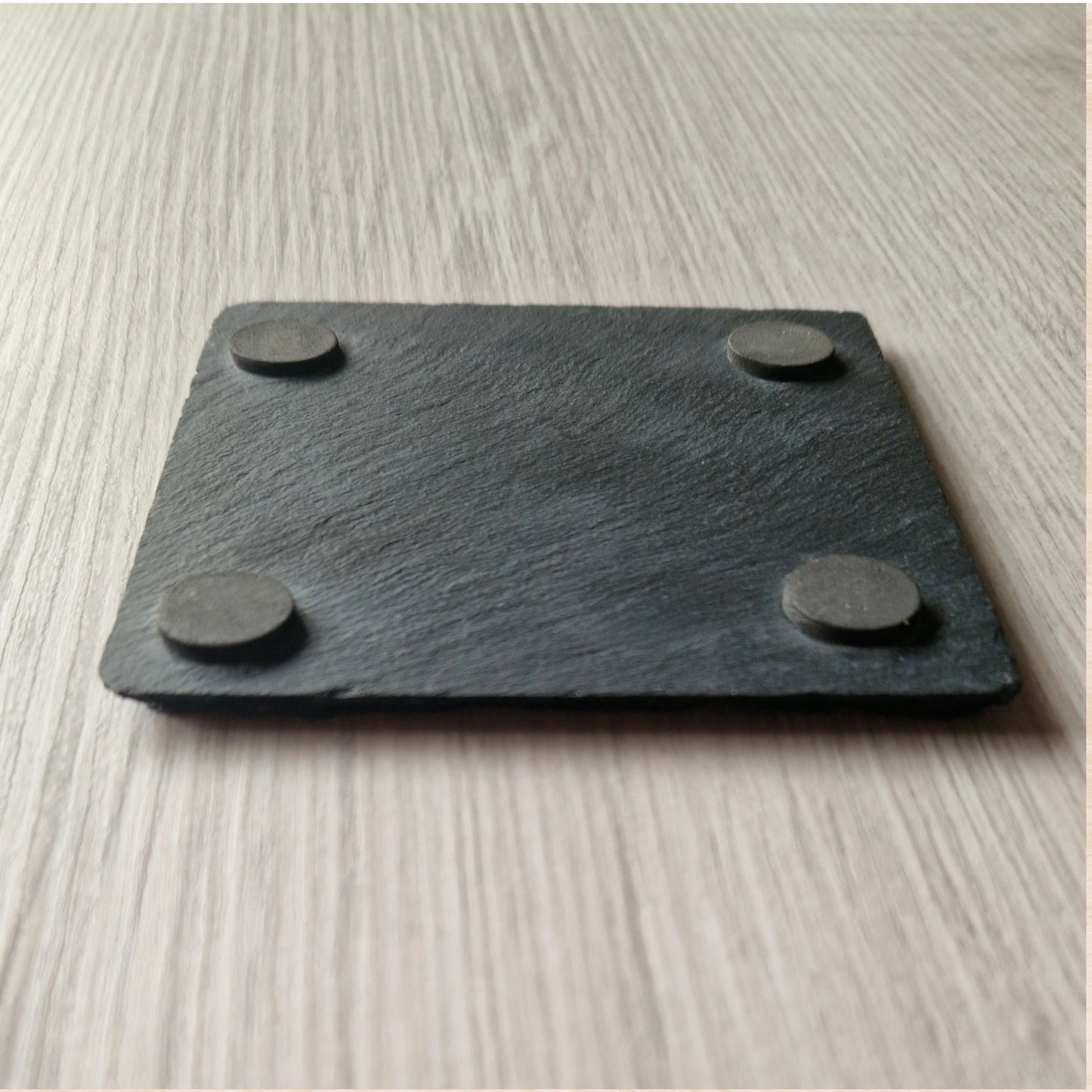 The underside of a black slate coaster, showing its natural rough texture. Each corner features a small cushioned foot, providing grip and protecting surfaces from scratches.