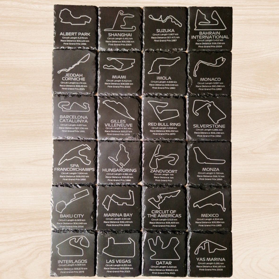 A set of 24 black slate coasters, each featuring an engraved design of a different F1 racetrack. The coasters showcase the outlines of each circuit, with additional engraved text listing the circuit length, race distance, and the year the track hosted its first Grand Prix. The intricate engravings are displayed against the natural texture of the slate, offering a stylish and informative collection of F1 race tracks