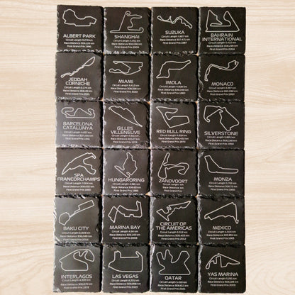 A set of 24 black slate coasters, each featuring an engraved design of a different F1 racetrack. The coasters showcase the outlines of each circuit, with additional engraved text listing the circuit length, race distance, and the year the track hosted its first Grand Prix. The intricate engravings are displayed against the natural texture of the slate, offering a stylish and informative collection of F1 race tracks