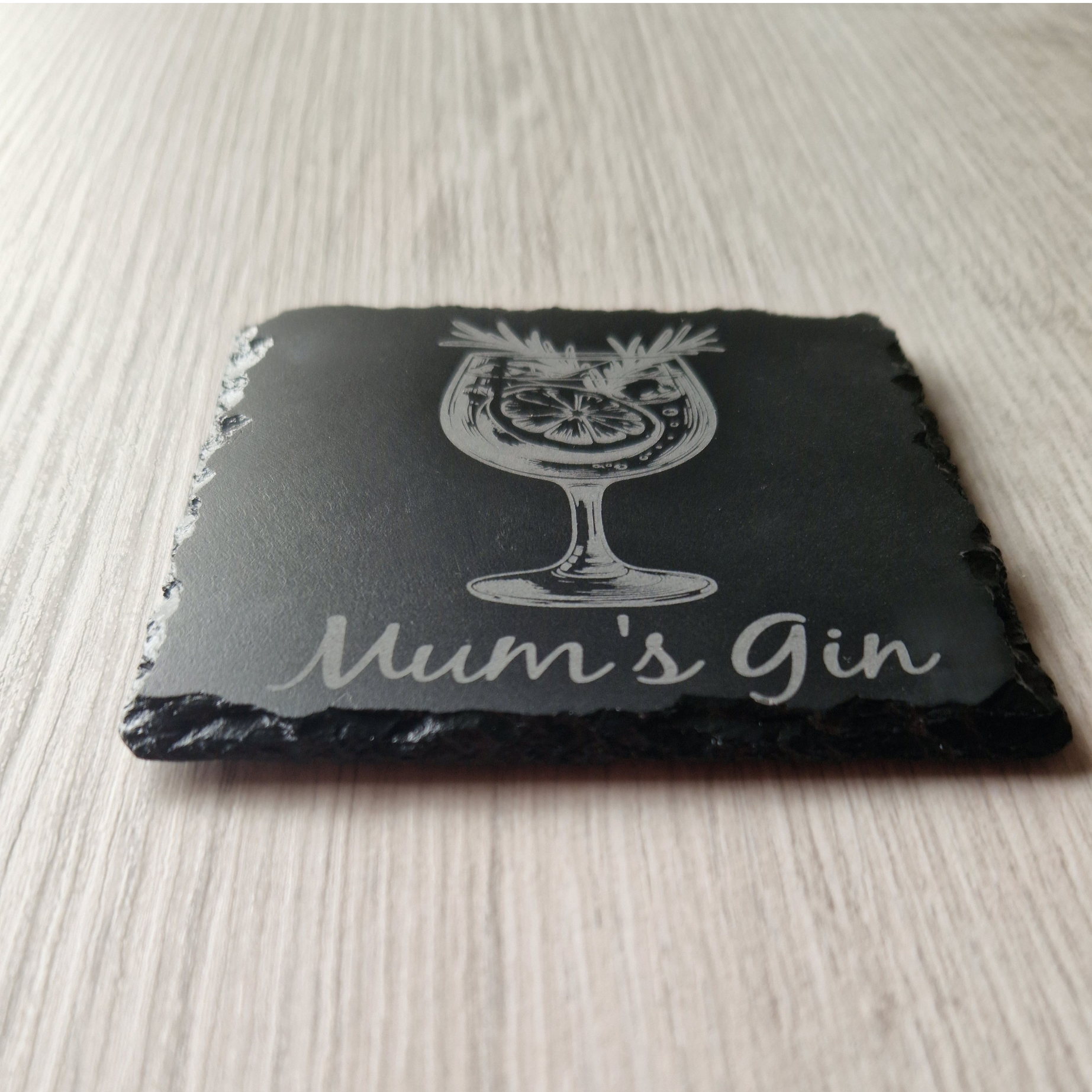 A close-up, low-angle view of a black slate coaster with an engraved gin glass and the words "Mum's Gin." The rough, natural texture of the slate's edges is visible, contrasting with the smooth, engraved surface on top.