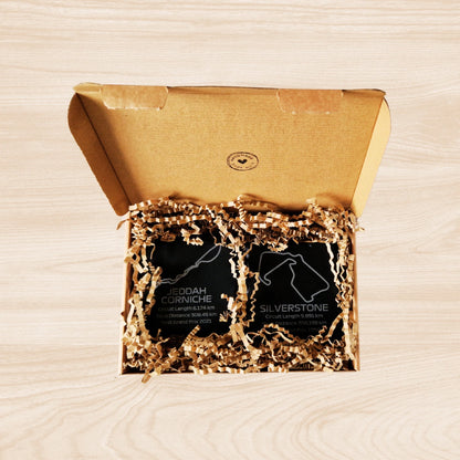 Two black slate racetrack coasters neatly arranged inside a cardboard shipping box. Each coaster features an engraved design of an F1 racetrack, with details such as the circuit length, race distance, and the year the track hosted its first Grand Prix. The coasters rest on a layer of protective material, and the box's interior emphasizes a secure, well-packed presentation. The natural texture of the slate contrasts with the smooth, precise engravings.