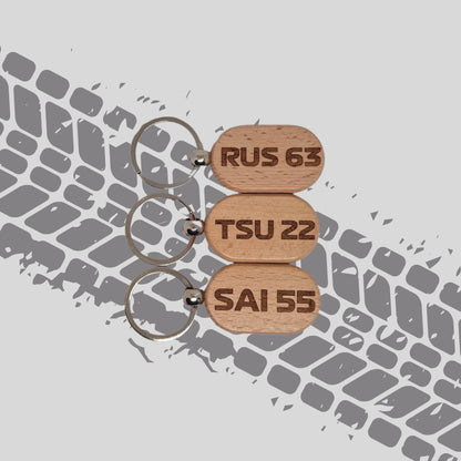 Three wooden keychains featuring F1 driver abbreviations and numbers for the 2025 season, displayed against a tyre tread background. Each keychain has a unique design with the driver's initials and number engraved on the wood