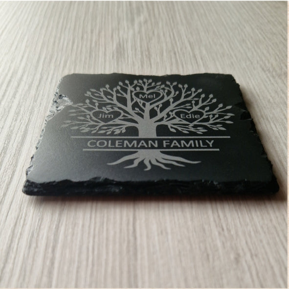 A close-up, low-angle view of a black slate coaster with an engraved family tree." The rough, natural texture of the slate's edges is visible, contrasting with the smooth, engraved surface on top.