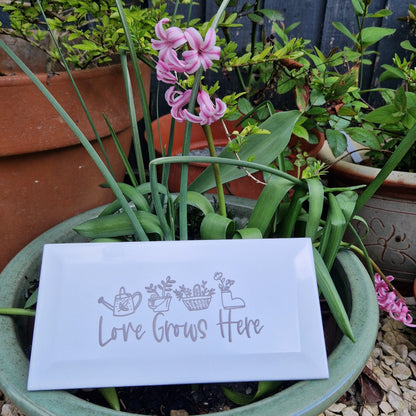 Engraved ceramic garden tile with 'Love Grows Here' placed outside next to a vibrant pink flower.