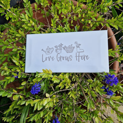 Engraved ceramic garden tile with 'Love Grows Here' displayed outdoors next to a green bush and purple flowers.