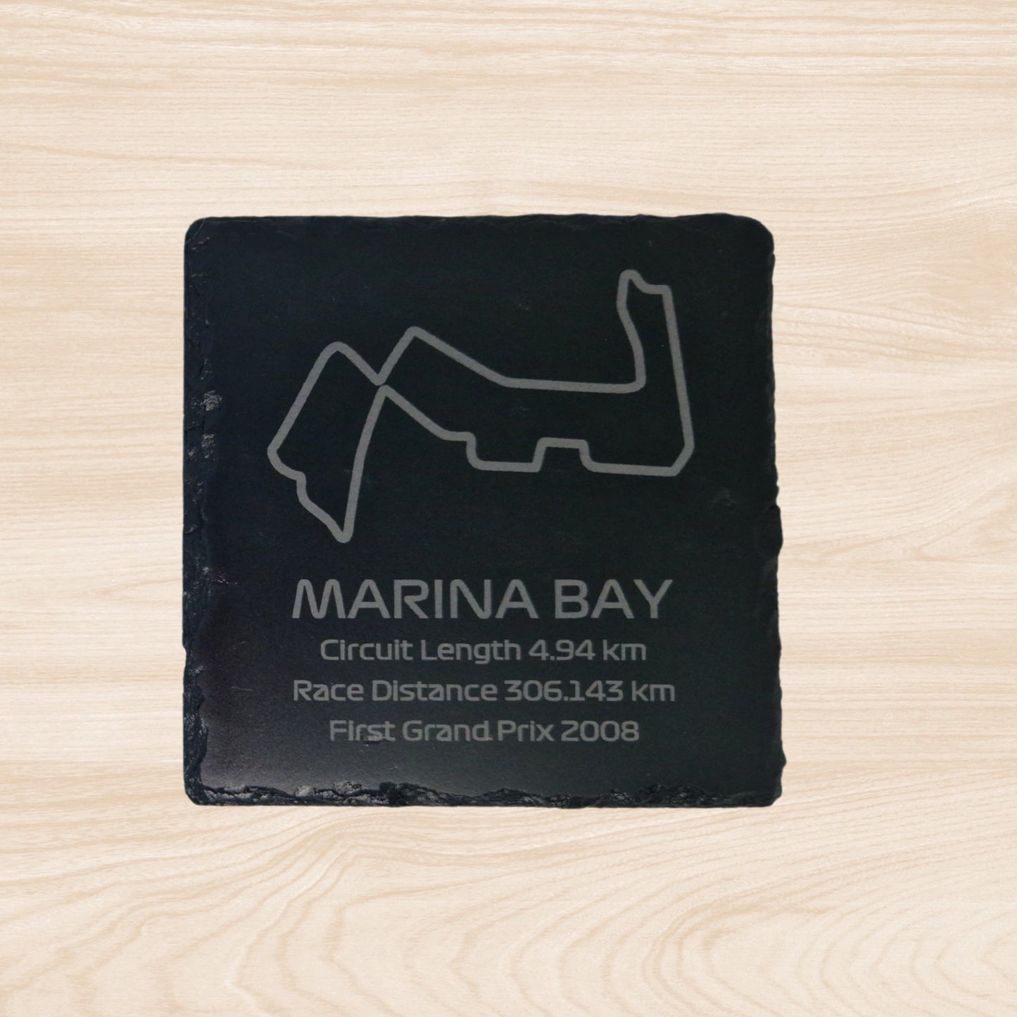 A black slate coaster featuring an engraved design of the Marina Bay racetrack. The engraving includes detailed outlines of the circuit, along with text listing the circuit length, race distance, and the year the track held its first Grand Prix. The sleek, engraved lines contrast with the natural rough texture of the slate, creating a unique, informative design.