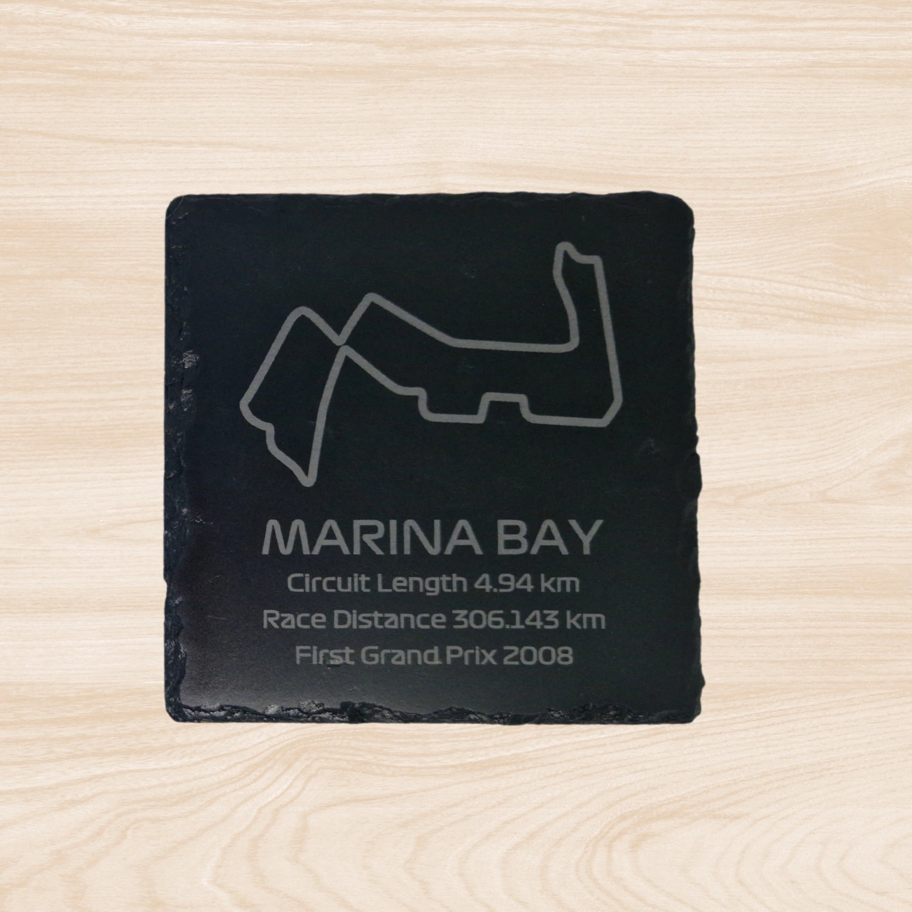 A black slate coaster featuring an engraved design of the Marina Bay racetrack. The engraving includes detailed outlines of the circuit, along with text listing the circuit length, race distance, and the year the track held its first Grand Prix. The sleek, engraved lines contrast with the natural rough texture of the slate, creating a unique, informative design.