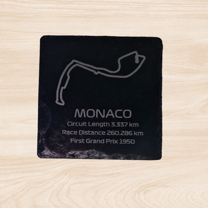 A black slate coaster featuring an engraved design of the Monaco racetrack. The engraving includes detailed outlines of the circuit, along with text listing the circuit length, race distance, and the year the track held its first Grand Prix. The sleek, engraved lines contrast with the natural rough texture of the slate, creating a unique, informative design.