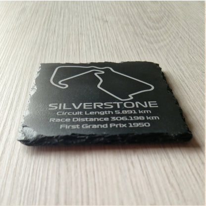 A close-up, low-angle view of a black slate coaster with an engraved outline of the Silverstone racetrack. The rough, natural texture of the slate's edges is visible, contrasting with the smooth, engraved surface on top.
