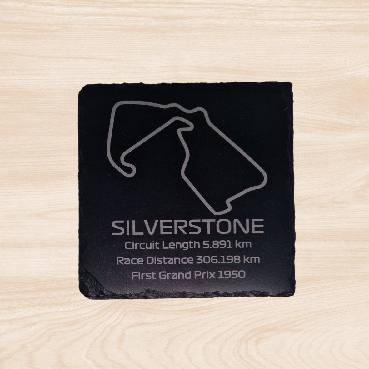 A black slate coaster featuring an engraved design of the Silverstone racetrack. The engraving includes detailed outlines of the circuit, along with text listing the circuit length, race distance, and the year the track held its first Grand Prix. The sleek, engraved lines contrast with the natural rough texture of the slate