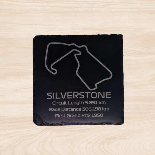 A black slate coaster featuring an engraved design of the Silverstone racetrack. The engraving includes detailed outlines of the circuit, along with text listing the circuit length, race distance, and the year the track held its first Grand Prix. The sleek, engraved lines contrast with the natural rough texture of the slate
