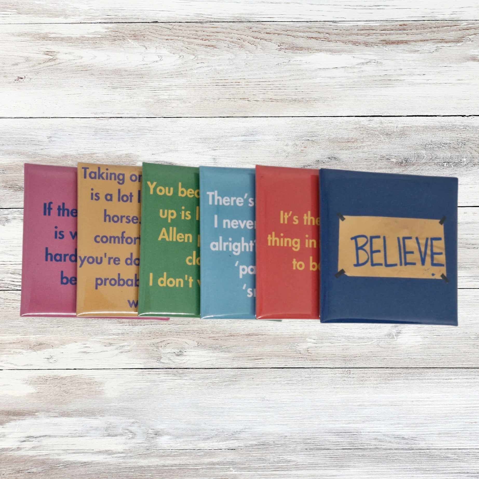 A set of six square fridge magnets inspired by Ted Lasso, featuring iconic quotes and designs from the show. They are in a line overlapping each other. The background is pale wood.