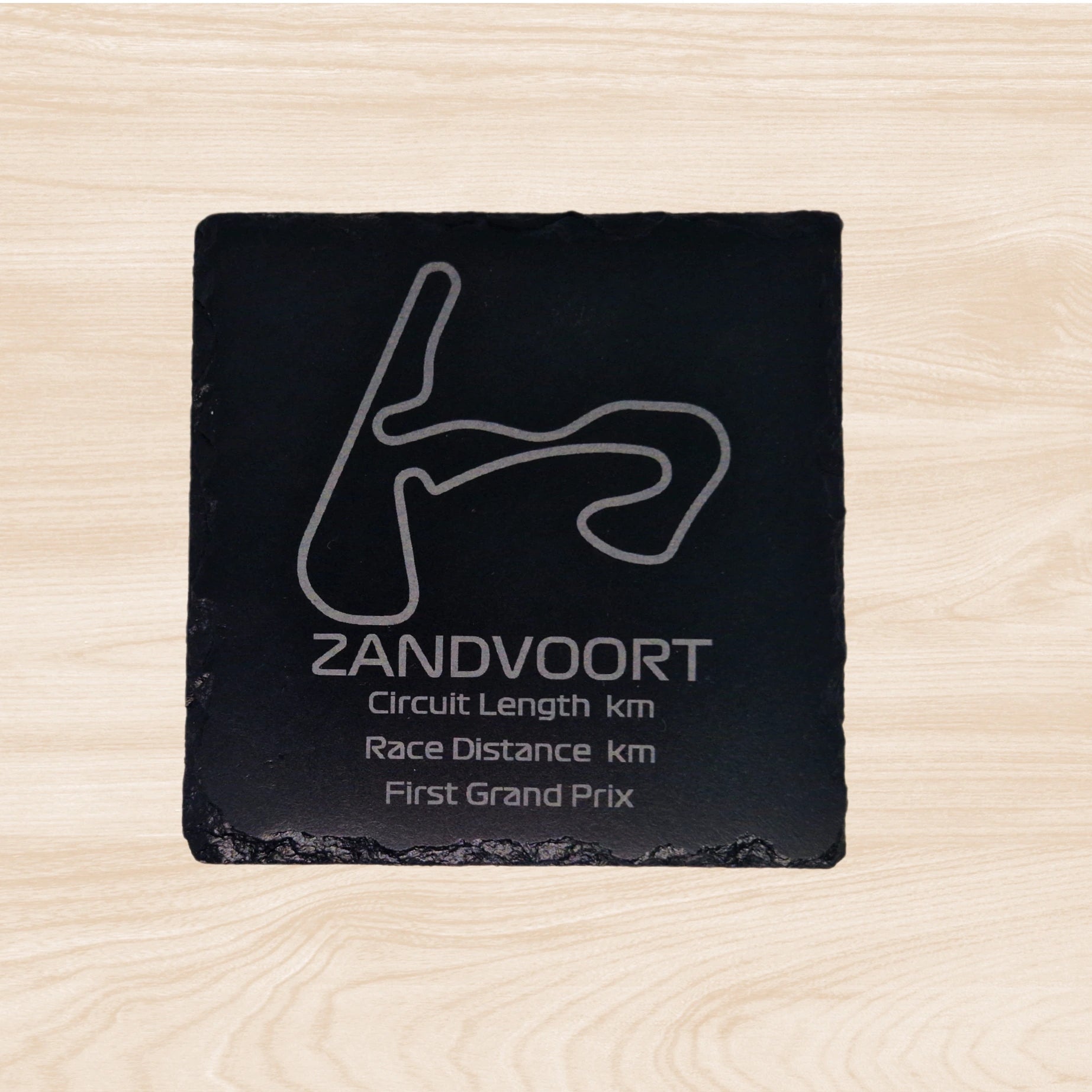 A black slate coaster featuring an engraved design of the Yas Marina racetrack. The engraving includes detailed outlines of the circuit, along with text listing the circuit length, race distance, and the year the track held its first Grand Prix. The sleek, engraved lines contrast with the natural rough texture of the slate, creating a unique, informative design.